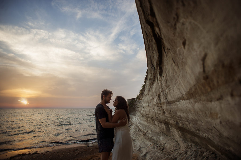 Greece Destination Engagement - Corfu - couple hugging next to a big rock, with sunset in background